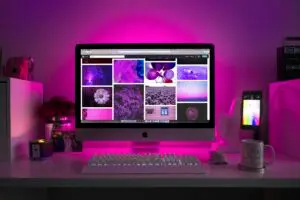 Creative workstation featuring an iMac and smartphone, displaying vibrant purple digital designs under pink ambient lighting.